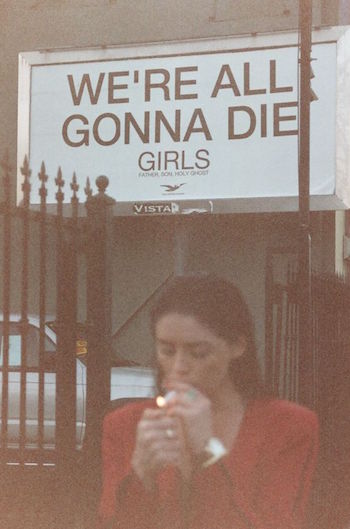 Girl Smoking Cigarette in Front Sign Saying We're All Gonna Die